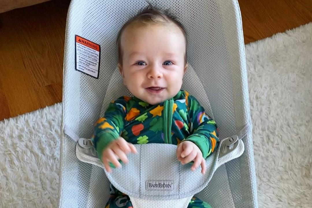 A smiling baby in a colorful floral outfit sits in a light blue baby bouncer, with a fuzzy rug and wooden floor in the background.