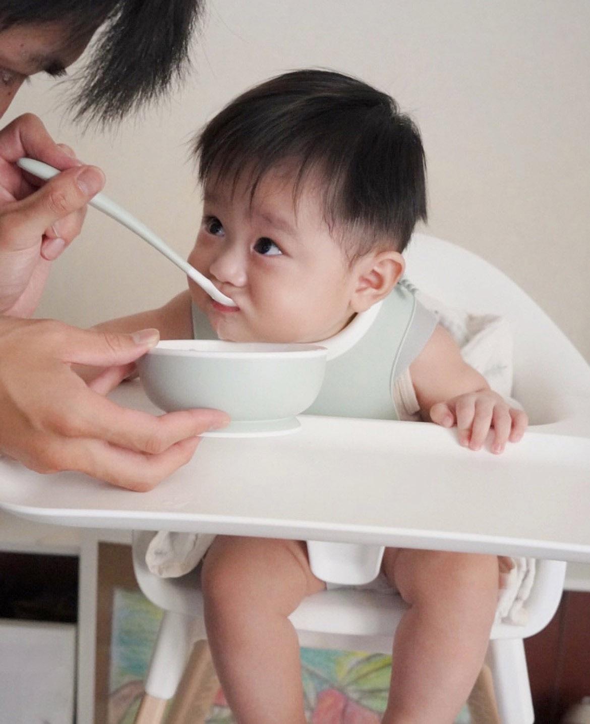 Baby sitting in a high chair being fed with a spoon by an adult. The baby is looking up towards the adult.