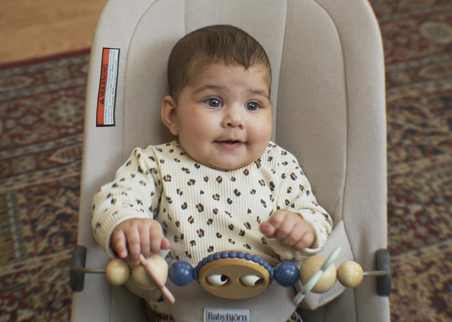 A baby is sitting in a beige bouncer wearing a white patterned outfit, holding onto a toy bar with wooden beads. A patterned rug is in the background.