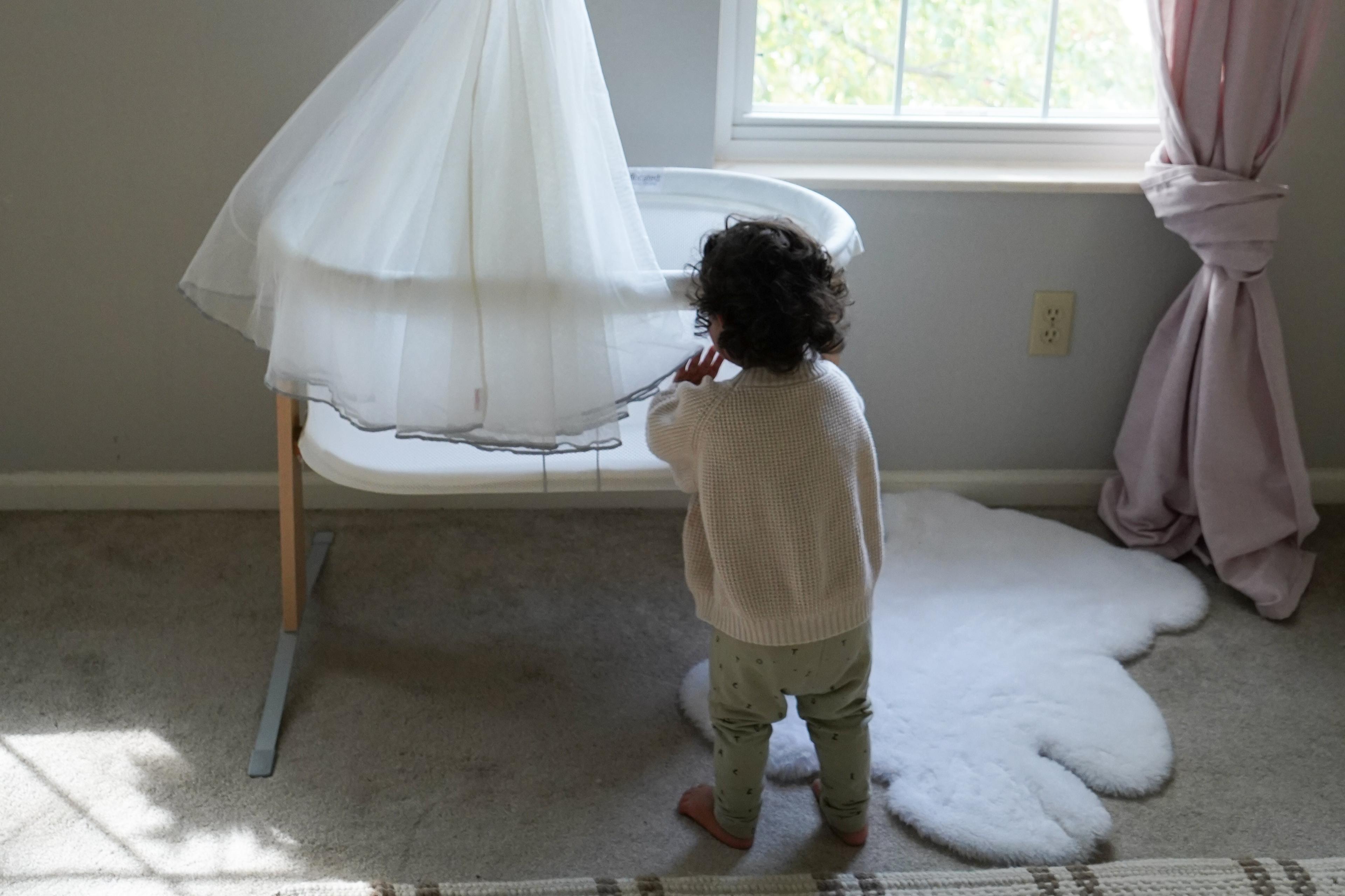 A toddler stands next to a white bassinet with a sheer canopy in a softly lit room, featuring a cloud-shaped rug and a window with pink curtains.