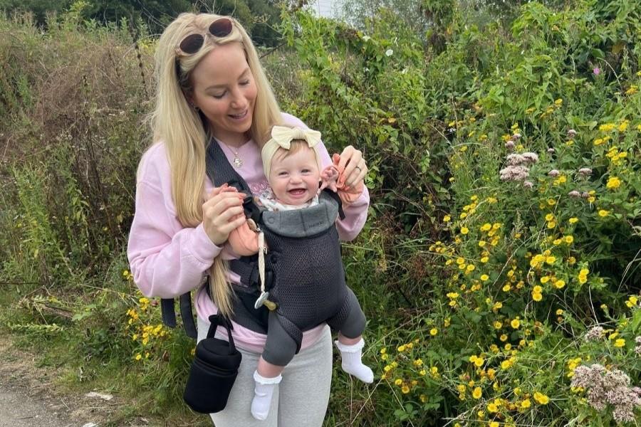 A woman in a pink sweater holds a smiling baby in a carrier while walking along a wildflower path.