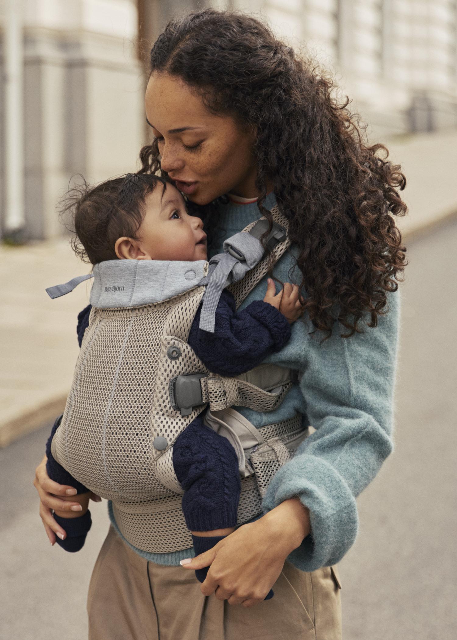 A woman with curly hair lovingly kisses a baby in a gray carrier. They are outdoors, with a building and street in the background.