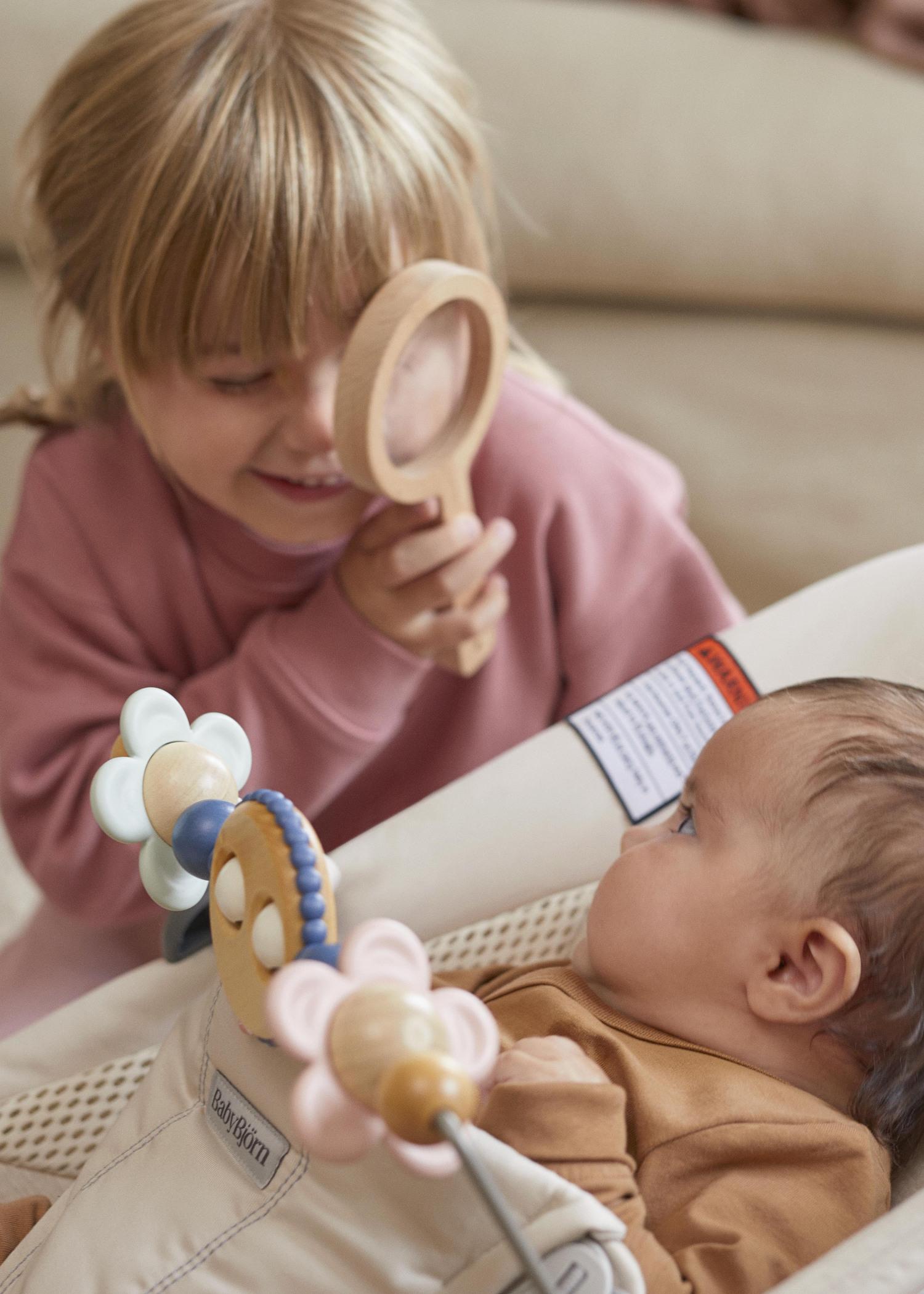 A child smiles while using a wooden magnifying glass to look at a baby lying in a bouncer with colorful toys attached.