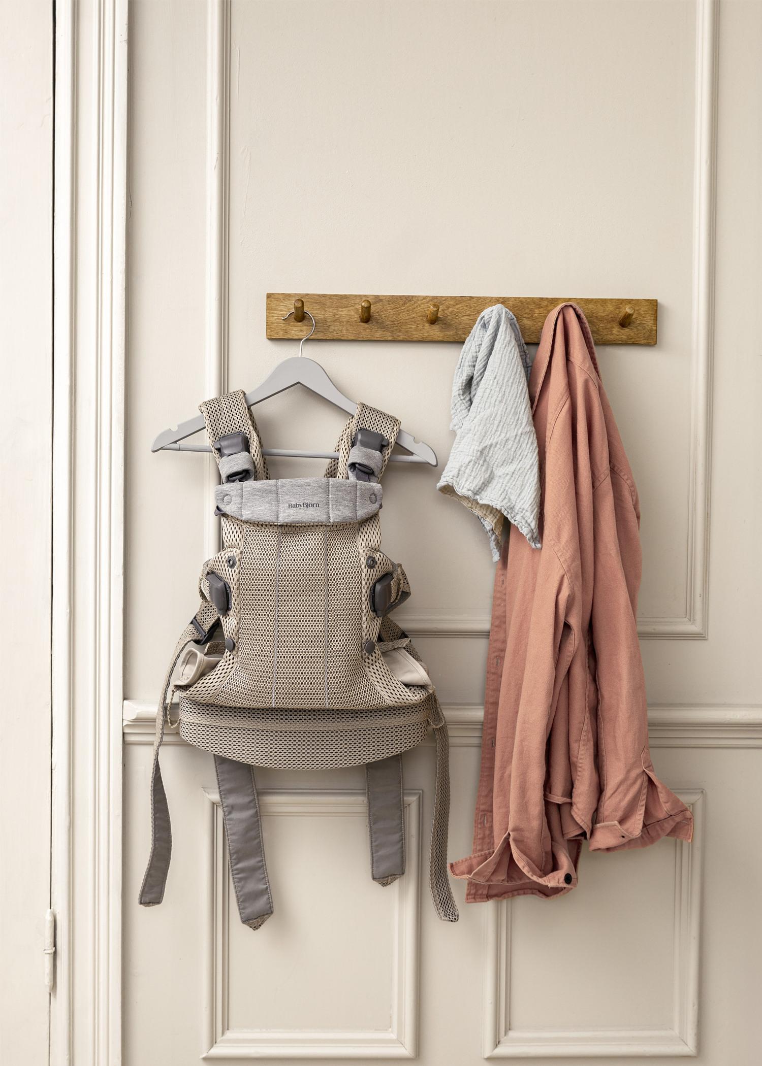 A beige baby carrier hangs on a wooden rack, alongside a light blue cloth and a pink shirt, against a cream-colored door.