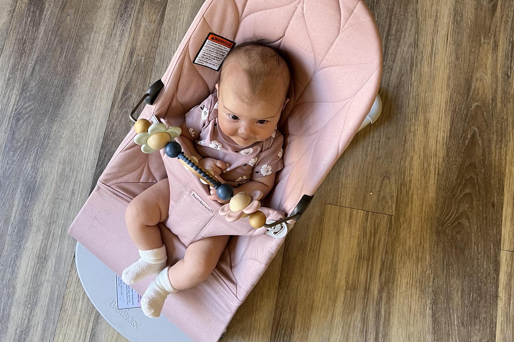 Baby in a pink bouncer with a toy bar, wearing a patterned onesie and white socks, on a wooden floor.