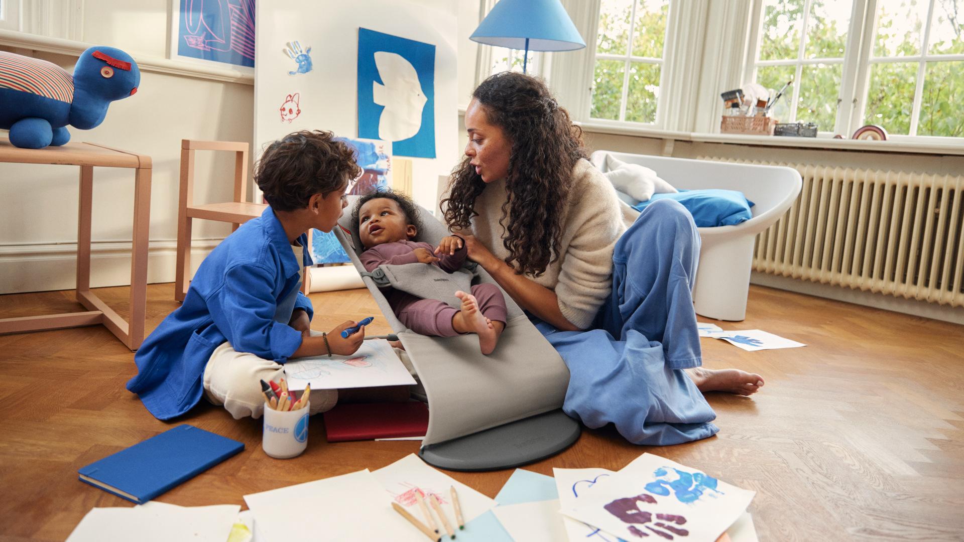 A woman and two children sit on a wooden floor with art supplies, surrounded by drawings. One child is in a gray baby carrier.